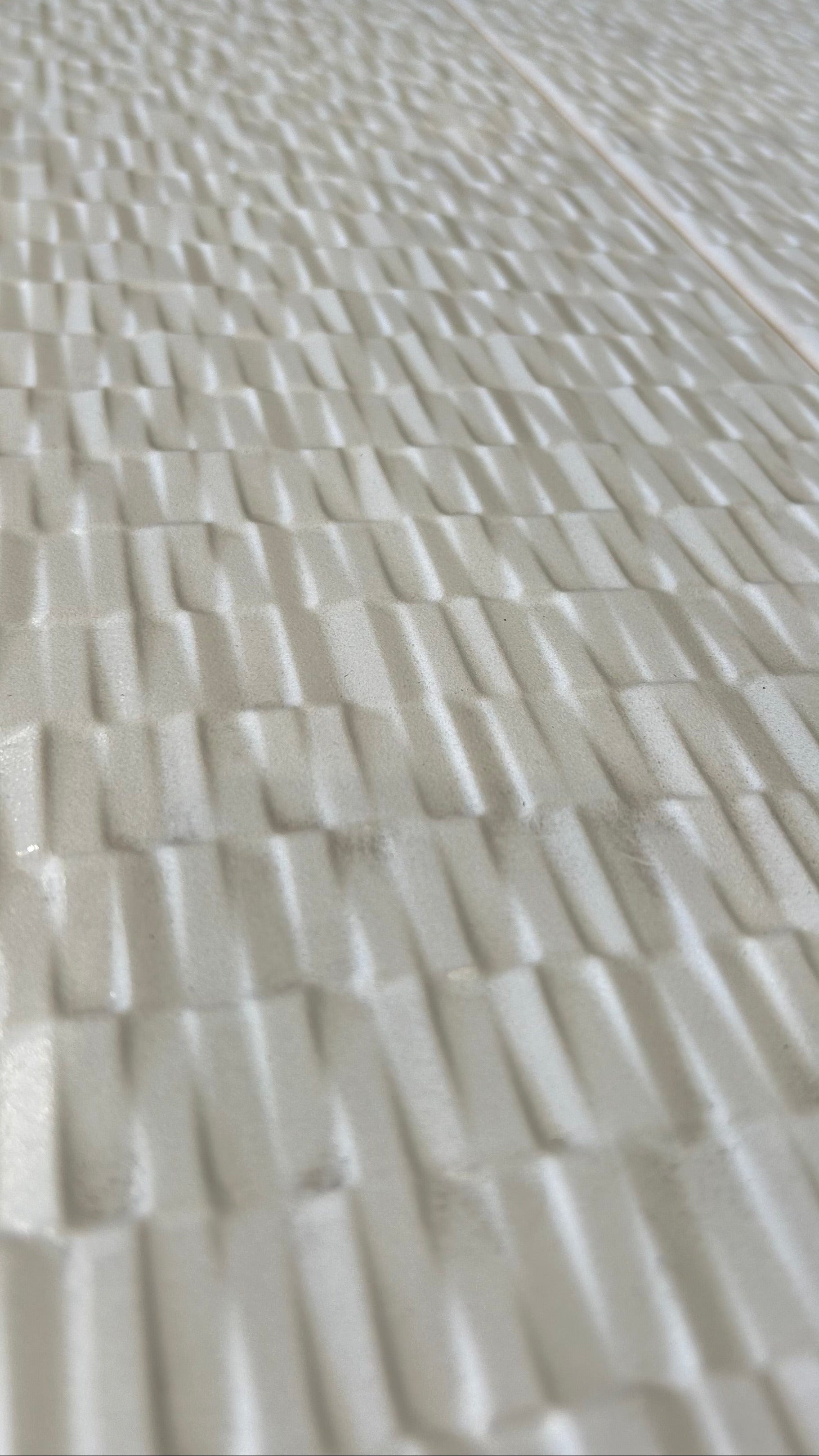 A close-up view of a textured, woven-look surface in warm off-white or cream, showing a repeating basketweave pattern white tile.